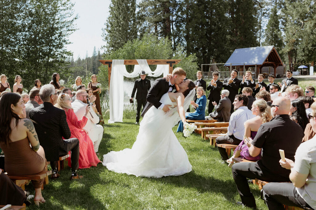 Wedding couple kissing in aisle after ceremony with guests on either side at Kinship Ranch with pine trees and greenery in background