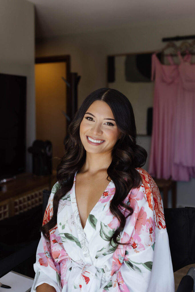 The bride smiles in a floral robe after finishing her hair and makeup for her Emerald Bay wedding