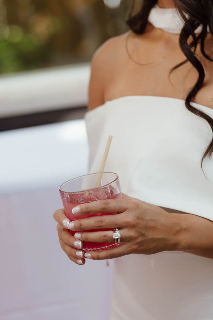 The bride holds a pink signature cocktail, showing her wedding rings and pearlescent manicure