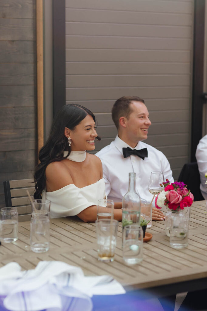 The bride and groom laugh together while listening to speeches at their wedding reception