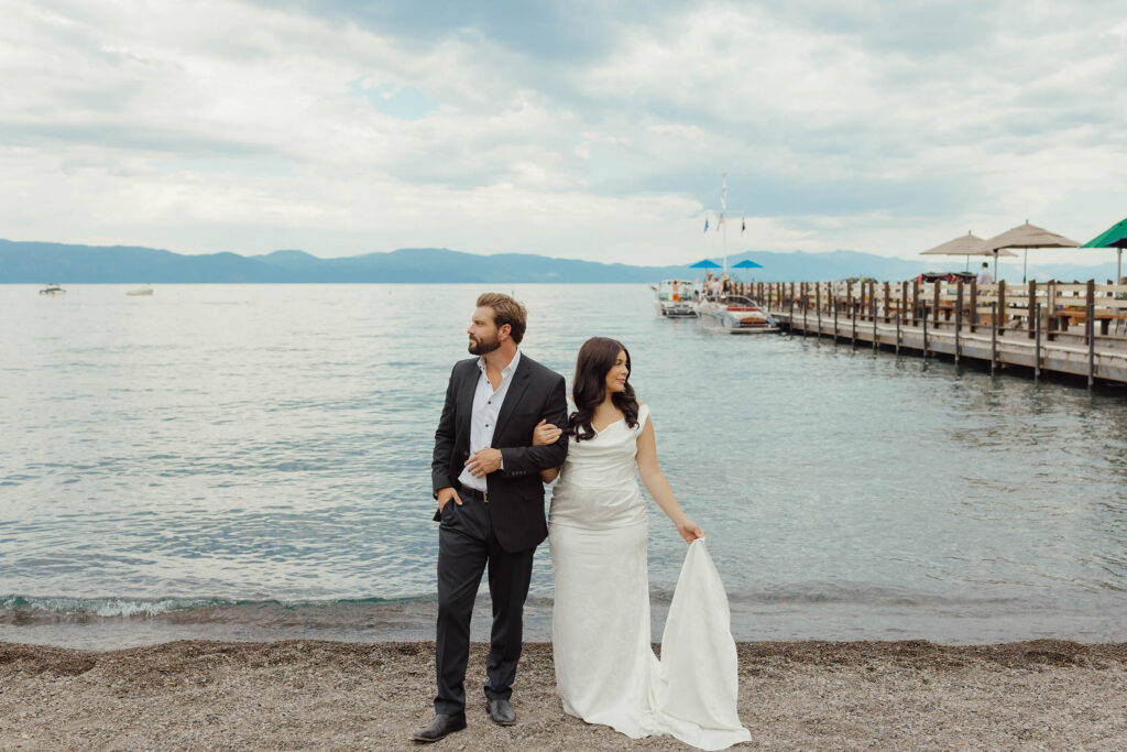 Bride and groom stand arm in arm along the Lake Tahoe shoreline looking in opposite directions
