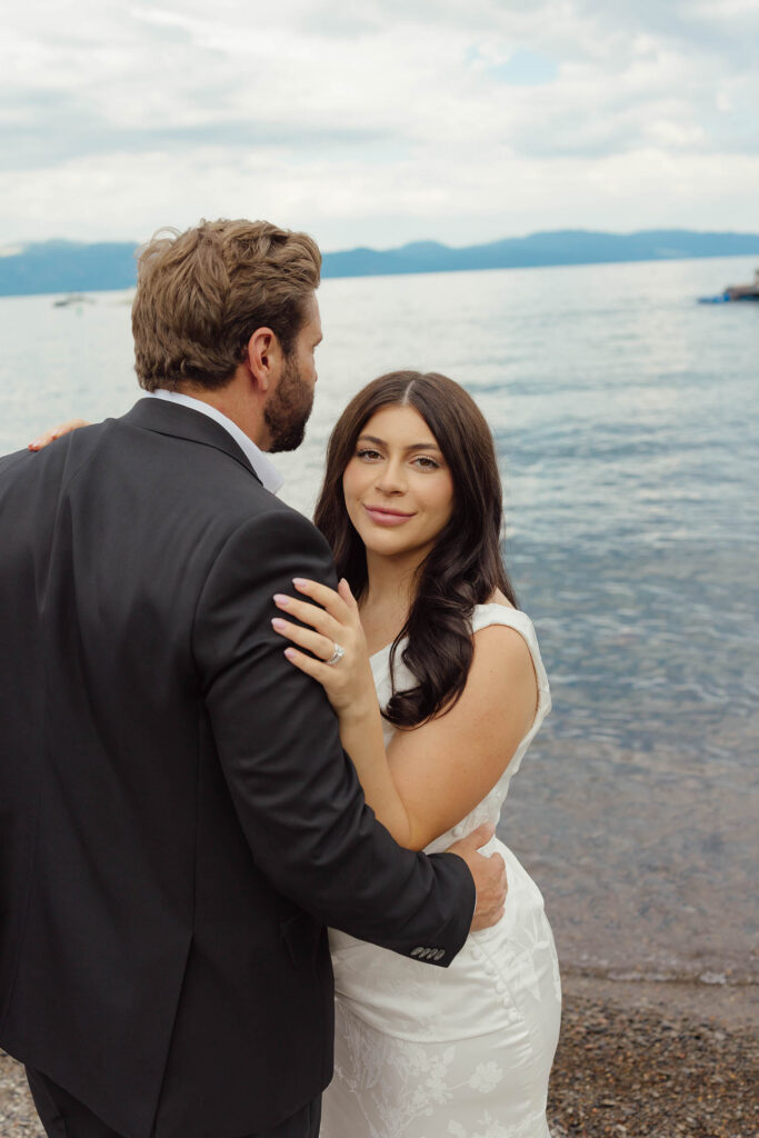 Bride hugging the groom beside Lake Tahoe, showing her wedding ring and smiling at the camera