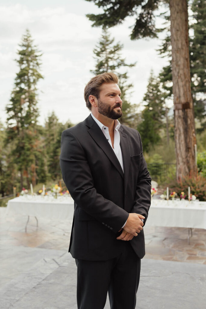 Groom waits for the ceremony in a black suit surrounded by tall pine trees