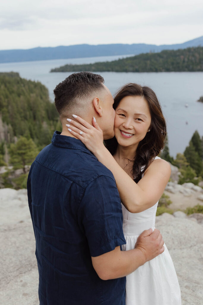Man kissing fiancé on cheek as she smiles at the camera and the couple holds each other at Emerald Bay