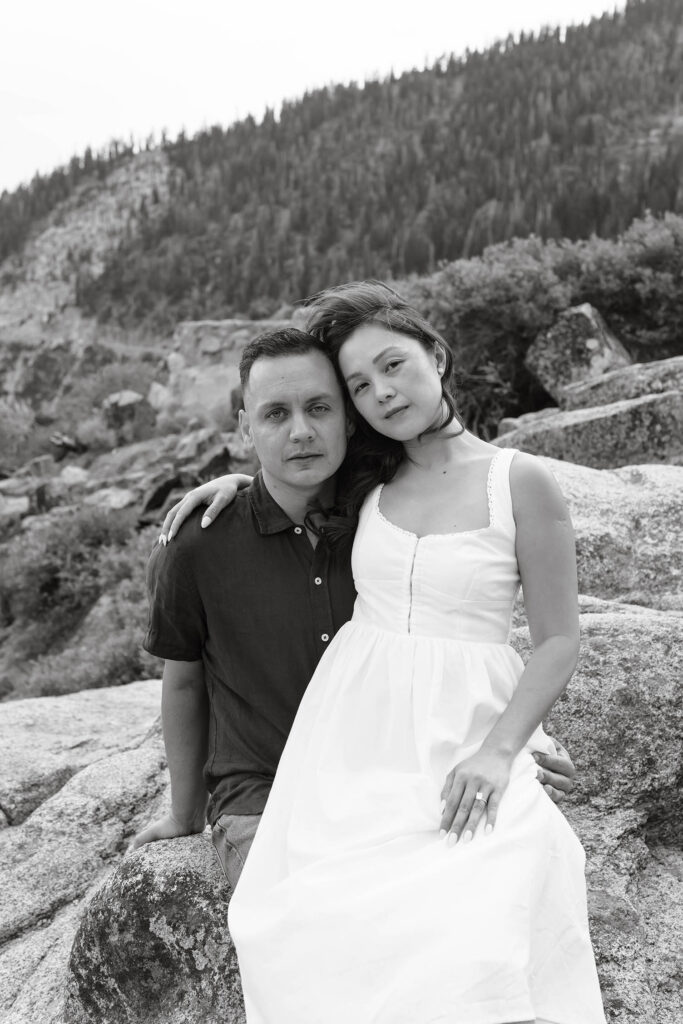 Engagement couple siting on large rock together while looking at camera at Emerald Bay with forest of pine trees in background