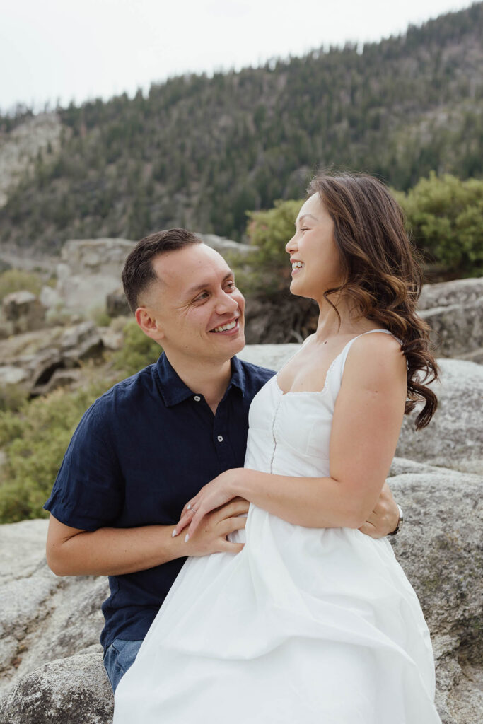 Engagement couple laughing while woman sits on fiancé's lap at Emerald Bay with pine trees in background