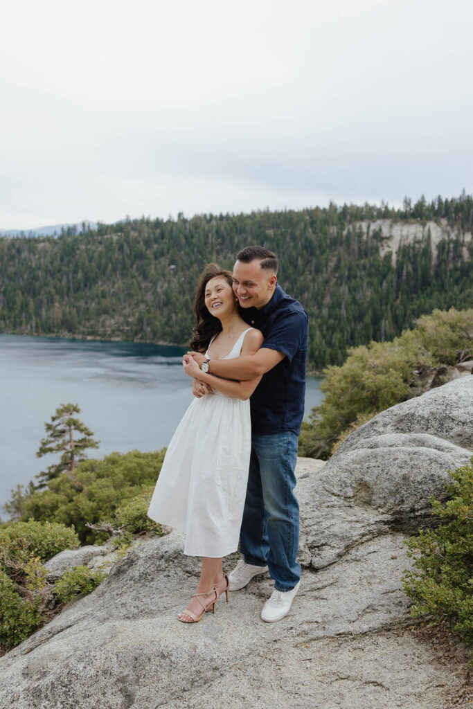 Man hugging fiancé from behind while standing on rocky ledge with Emerald Bay in background