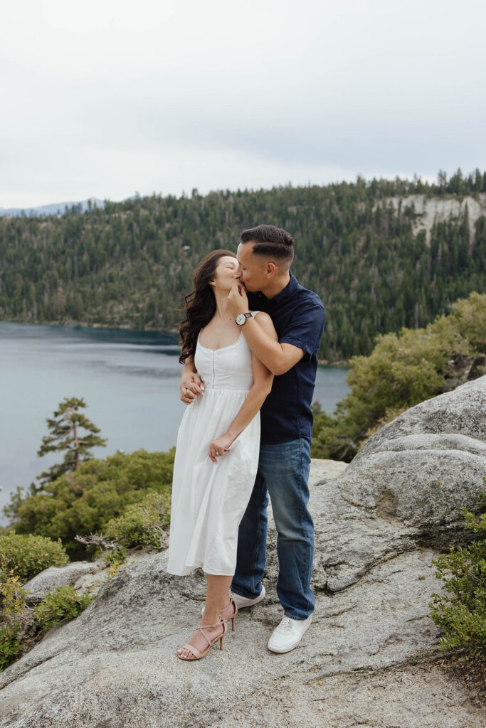 Engagement couple kissing while holding hands and standing on rocky ledge at Emerald Bay