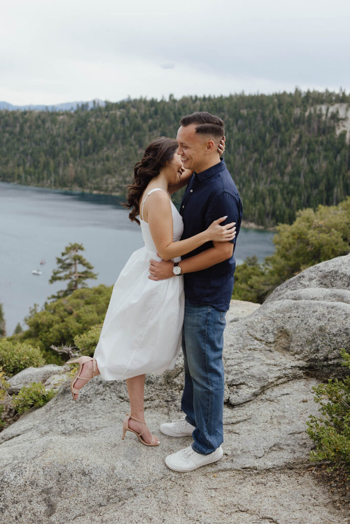 Engagement couple hugging on rocky ledge while smiling with Emerald Bay in background