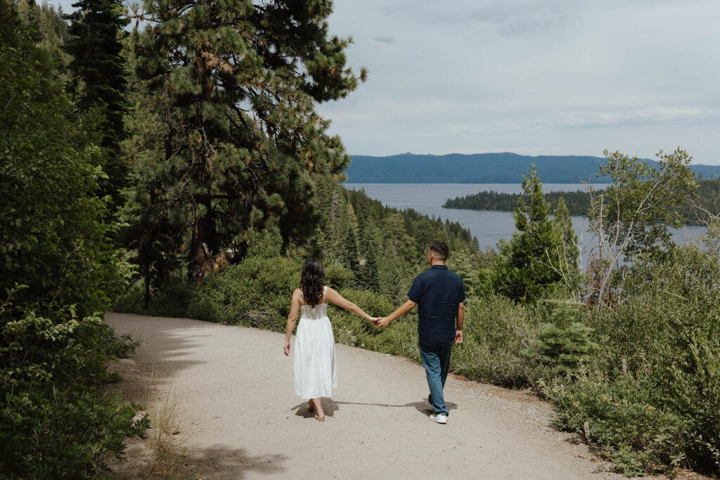 Engagement couple holding hands while walking along dirt trail together next to pine trees and greenery at Emerald Bay