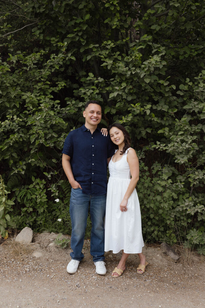 Engagement couple standing next to each other and smiling at camera in front of lots of greenery at Emerald Bay