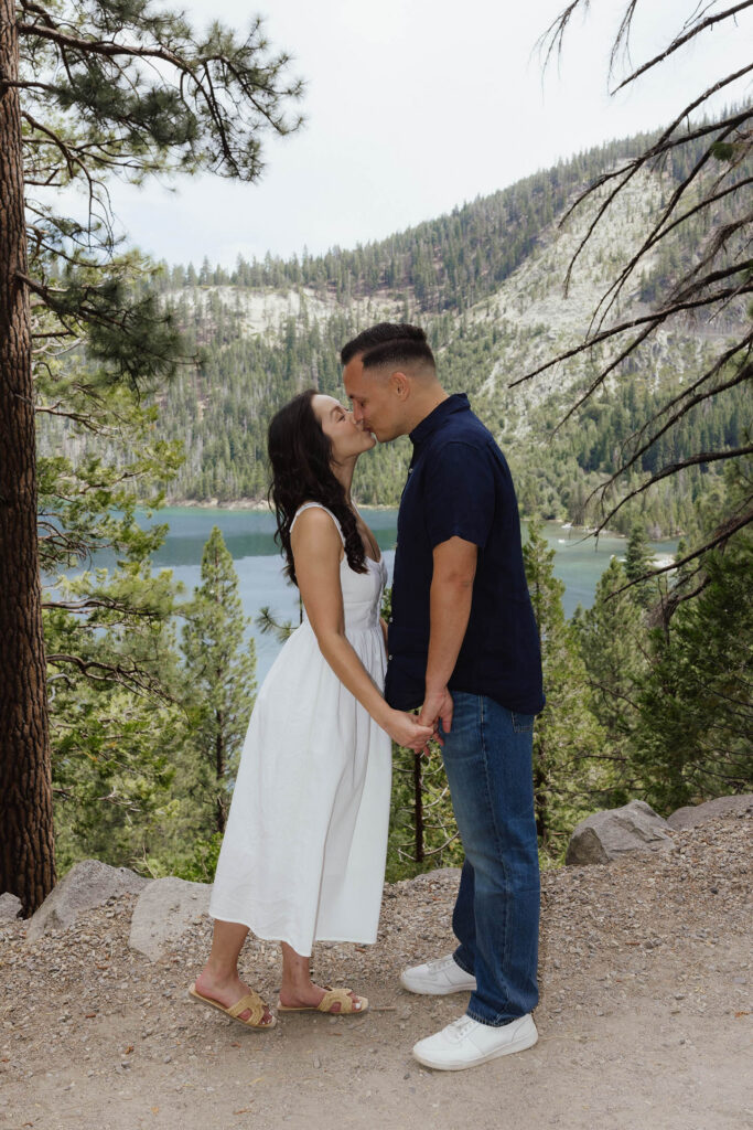 Engagement couple holding hands and kissing while on dirt trail with tall pine trees and Emerald Bay behind them
