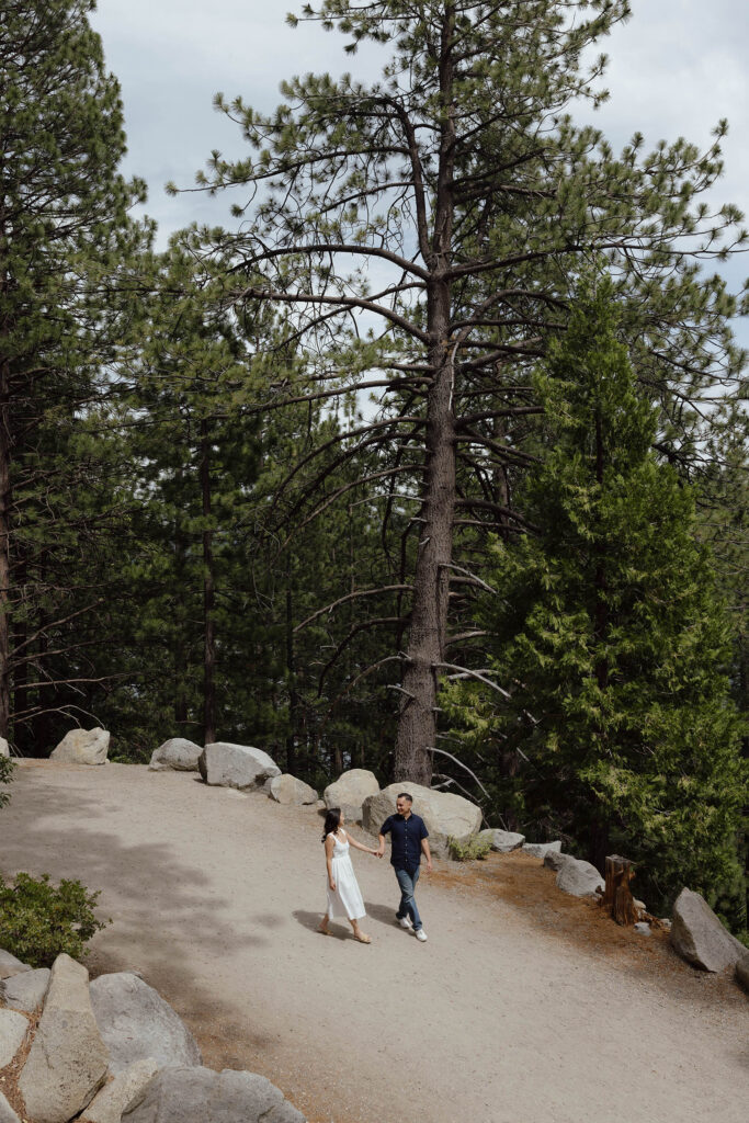 Engagement couple in the distance holding hands and walking along dirt trail next to large rocks and pine trees at Emerald Bay