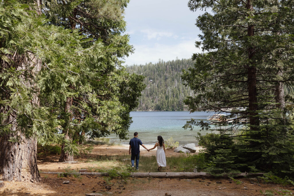 Engagement couple holding hands and walking through clearing together towards Emerald Bay with pine trees on either side of them