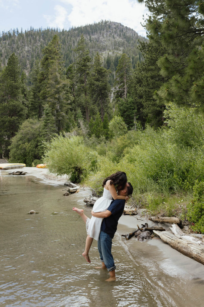 Man lifting fiancé up while couple kisses standing in water on a beach in Emerald Bay with lots of greenery in background