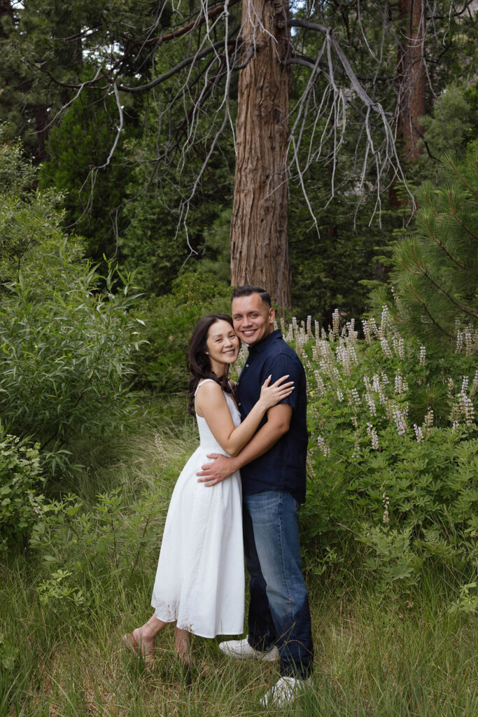 Engagement couple holding each other and smiling at camera while standing in front of pine trees and greenery at Emerald Bay
