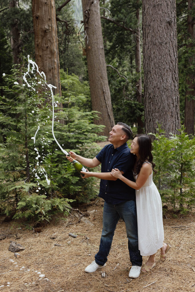 Engagement couple popping champagne together while surrounded by pine trees and greenery at Emerald Bay