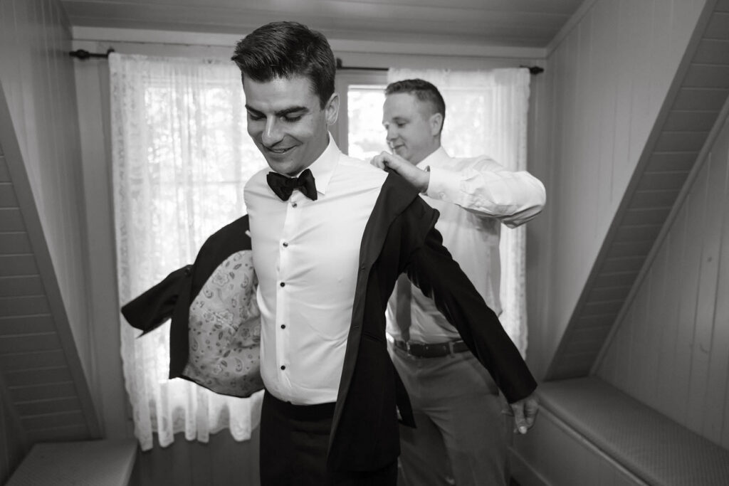 Black-and-white photo of a groomsman helping the groom into his tuxedo jacket