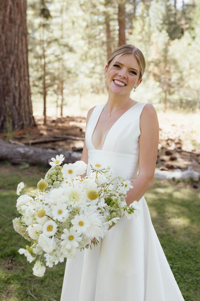 Smiling bride holds an oversized white wildflower bouquet beneath the pine trees