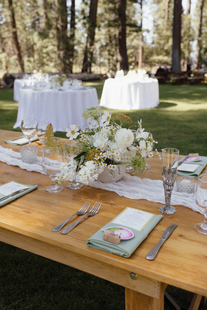 Wooden reception table decorated with white wildflowers, green napkins, and vintage glassware