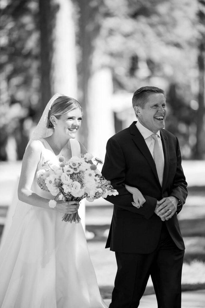 Black-and-white photo of the smiling bride walking with her father toward the ceremony