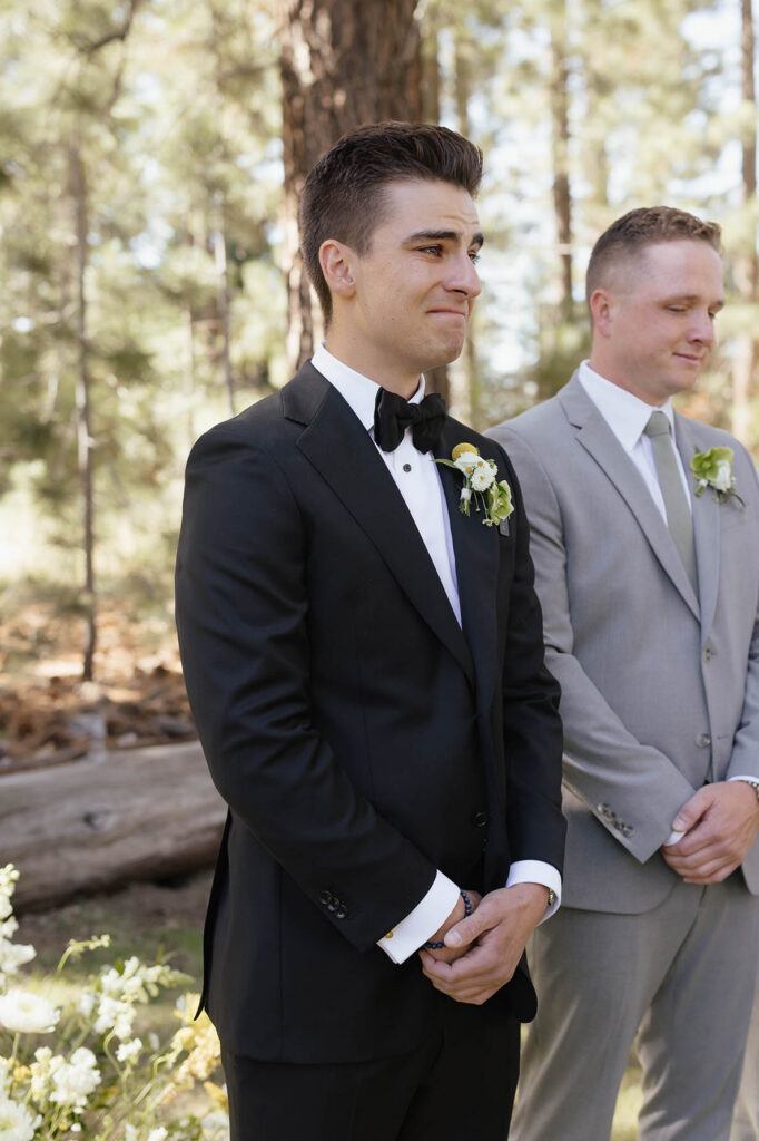 Groom holds back tears as he watches the bride approach through the pine trees