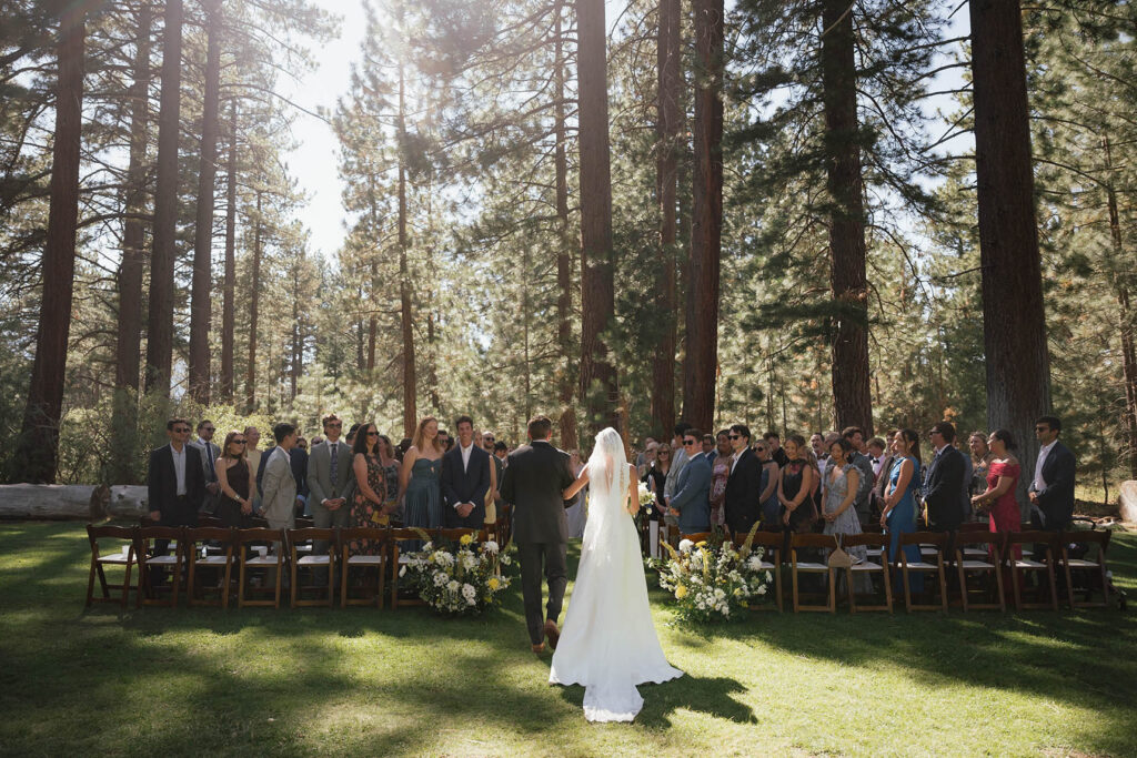 Bride and her father enter the forest ceremony surrounded by standing wedding guests