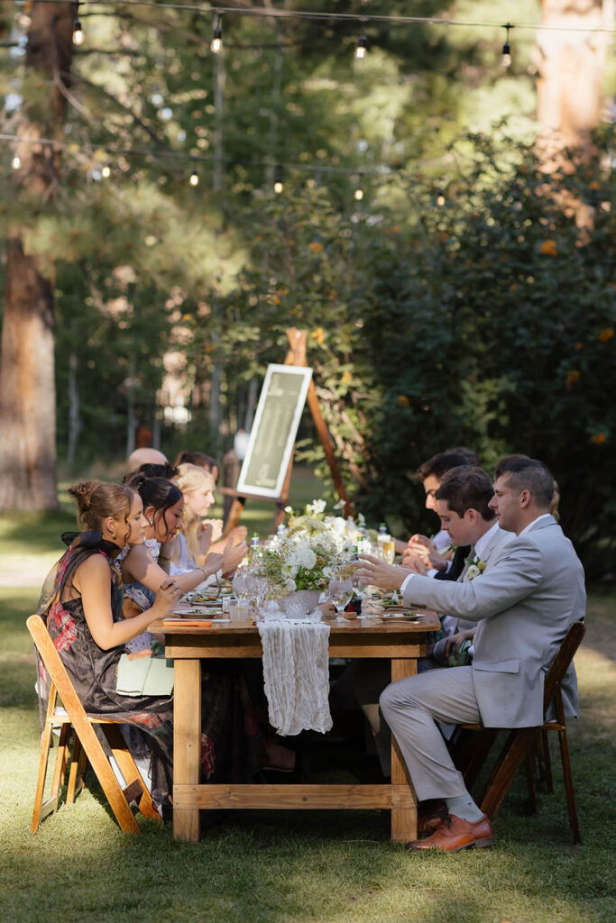 Wedding guests dine together at a long wooden table beneath the pine trees