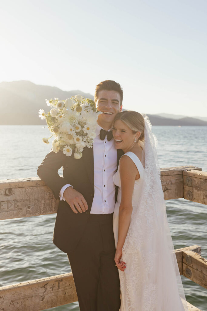 Laughing newlyweds leaning against the dock in Lake Tahoe with the bride’s white bouquet behind his shoulder