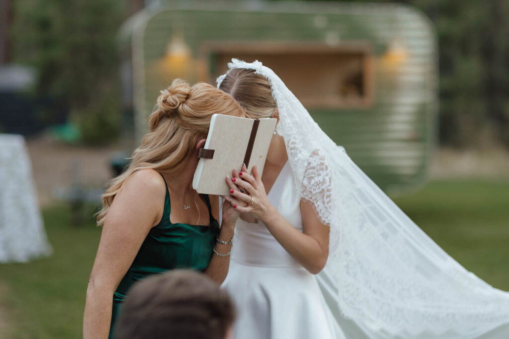 Bride and bridesmaid share a private laugh behind a vow book during the outdoor reception