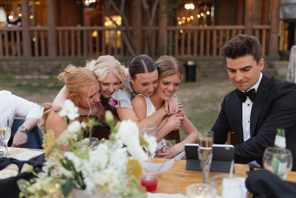 Friends embrace the smiling bride as the groom watches a video message beside her