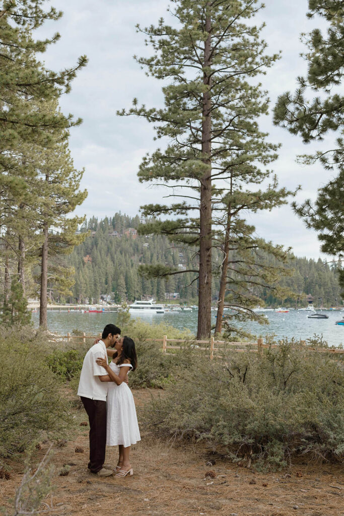 Engaged couple kissing on a forest trail with the lake and boats behind them