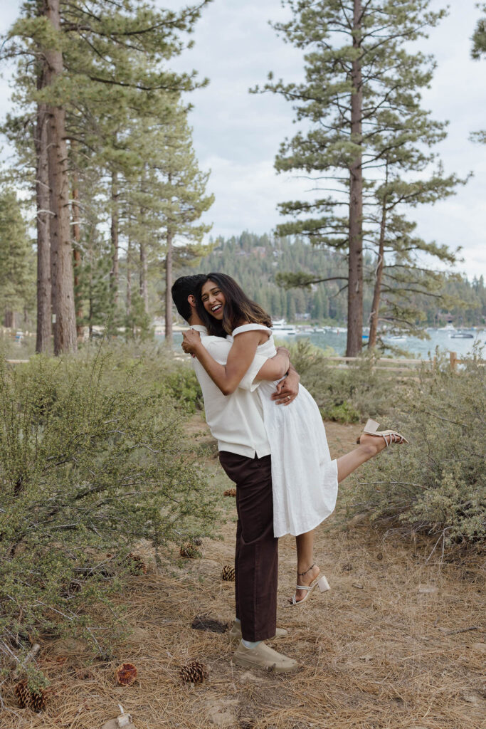 Laughing engaged couple hugging as he lifts her beside the Lake Tahoe shoreline