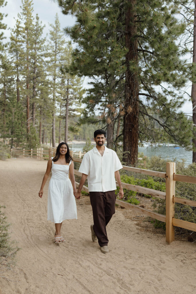 Engaged couple walking hand smiling in hand along a wooded trail beside Lake Tahoe