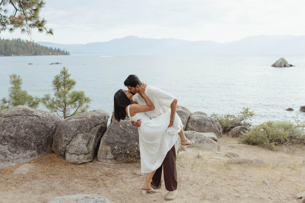 Man dipping and kissing his fiancée on a rocky overlook above Lake Tahoe