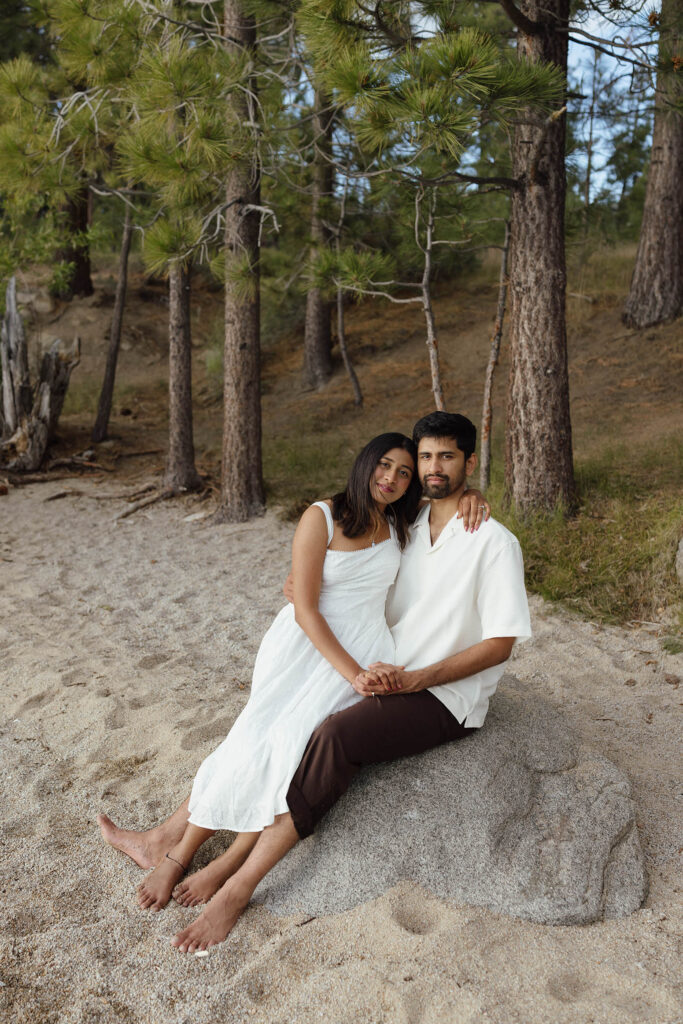 Barefoot engaged couple sitting together on a boulder beneath Lake Tahoe pine trees