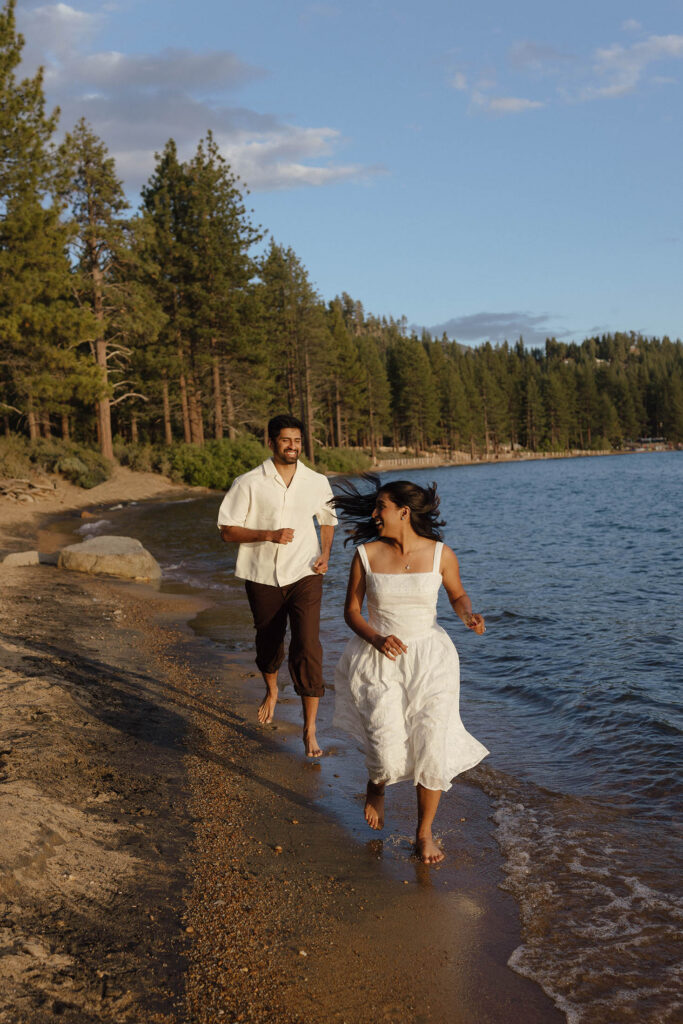 Laughing engaged couple running barefoot along the Lake Tahoe shoreline at golden hour