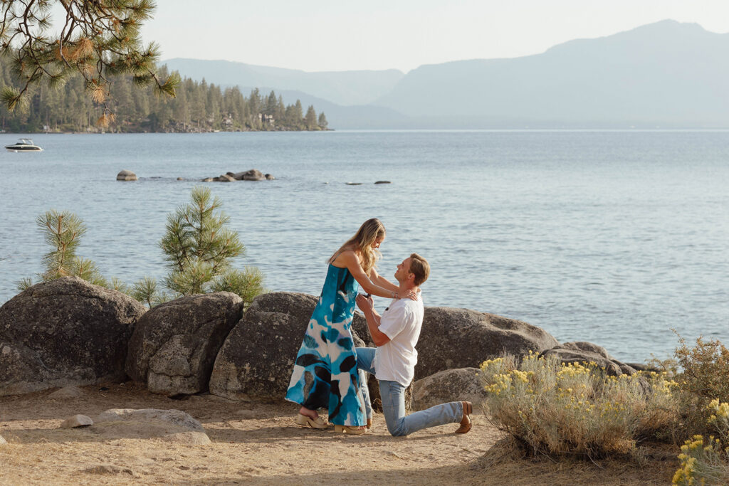 Man kneeling to propose to a woman in a blue patterned dress beside Lake Tahoe, surrounded by granite boulders and pines
