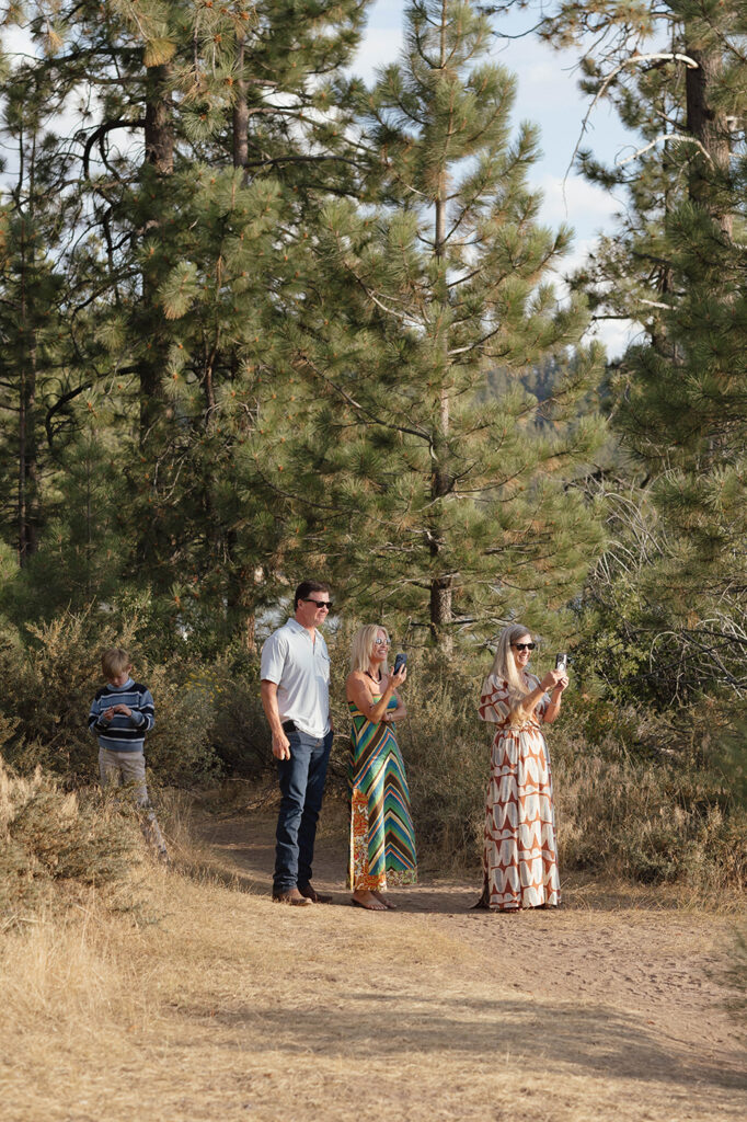 Family watching the proposal from a pine-lined trail, with two women holding up phones to capture the moment