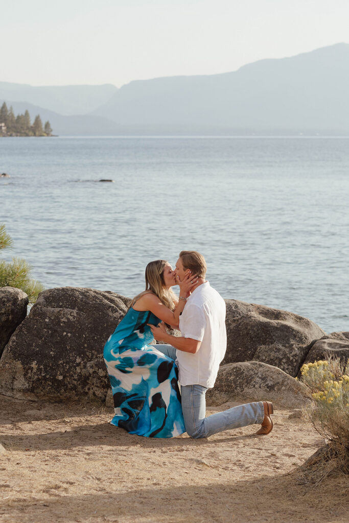 Couple kissing beside Lake Tahoe while the man remains on one knee and the woman cups his face