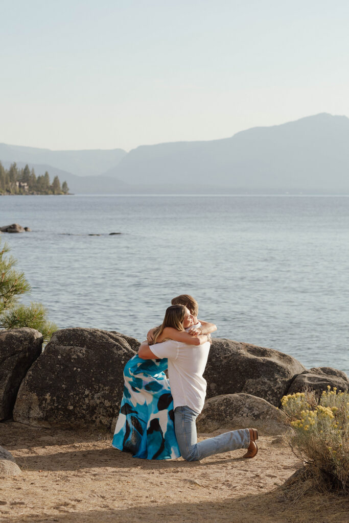Woman leaning down to embrace her kneeling partner after his proposal, with Lake Tahoe and mountains behind them