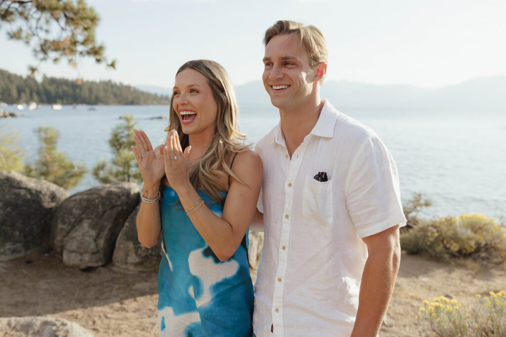 Newly engaged woman smiling with her hands raised and engagement ring visible as her partner stands beside her
