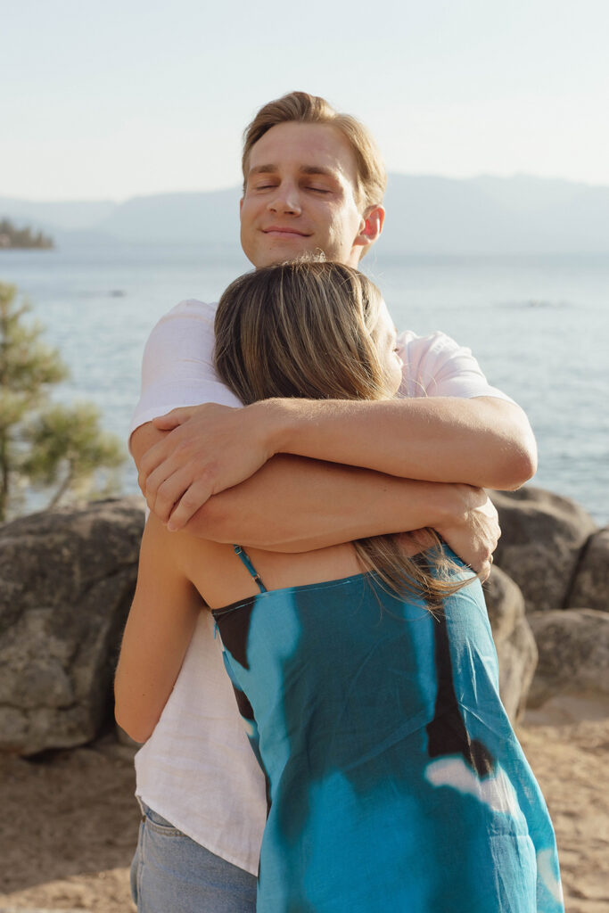 Newly engaged man closing his eyes and smiling as he hugs his fiancée tightly, with Lake Tahoe and granite boulders behind them