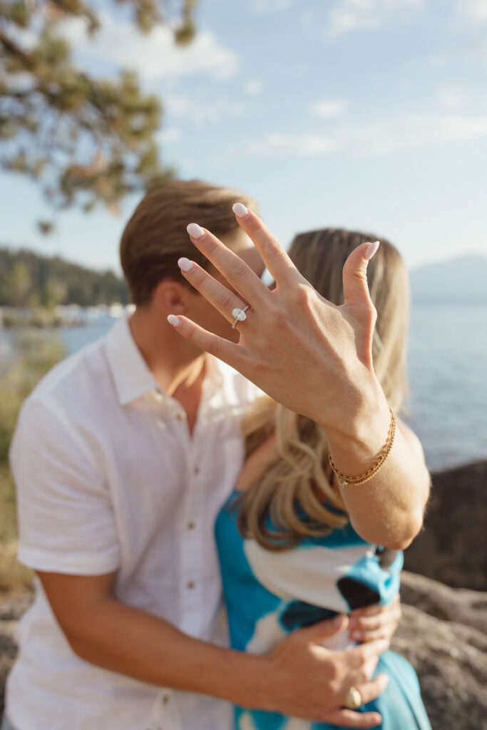 Woman holding her engagement ring toward the camera while kissing her fiancé beside Lake Tahoe