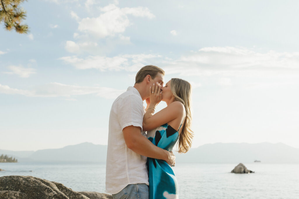 Newly engaged couple kissing along the Lake Tahoe shoreline as the woman cups her fiancé’s face