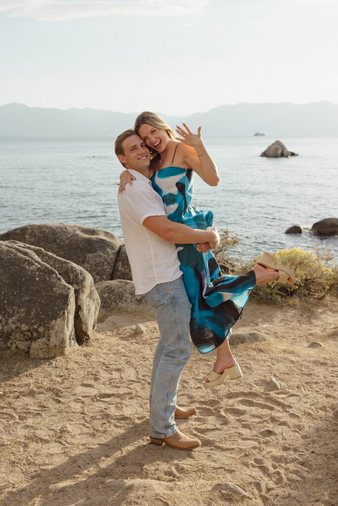 Man lifting his smiling fiancée as she shows her engagement ring and kicks one leg back beside Lake Tahoe
