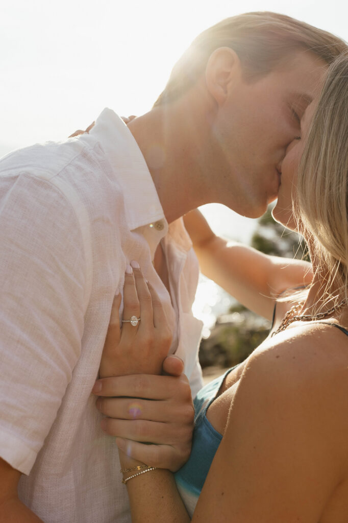 Sunlit close-up of the couple kissing, with her engagement ring visible as her hand rests against his white shirt