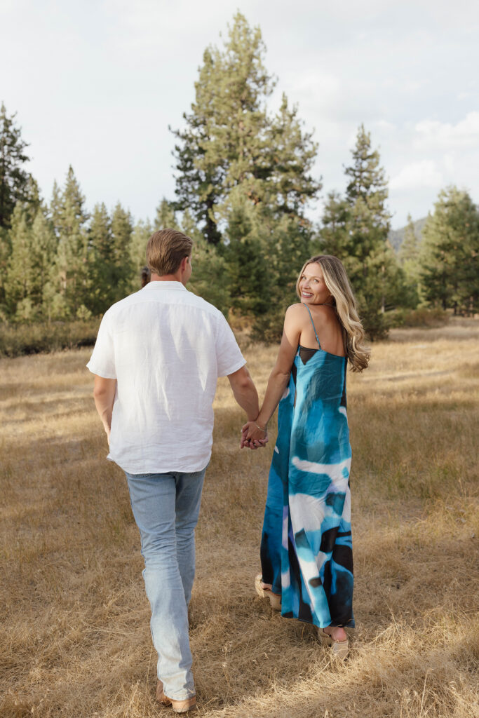 Couple walking hand in hand through golden grass and pine trees as the woman smiles back over her shoulder