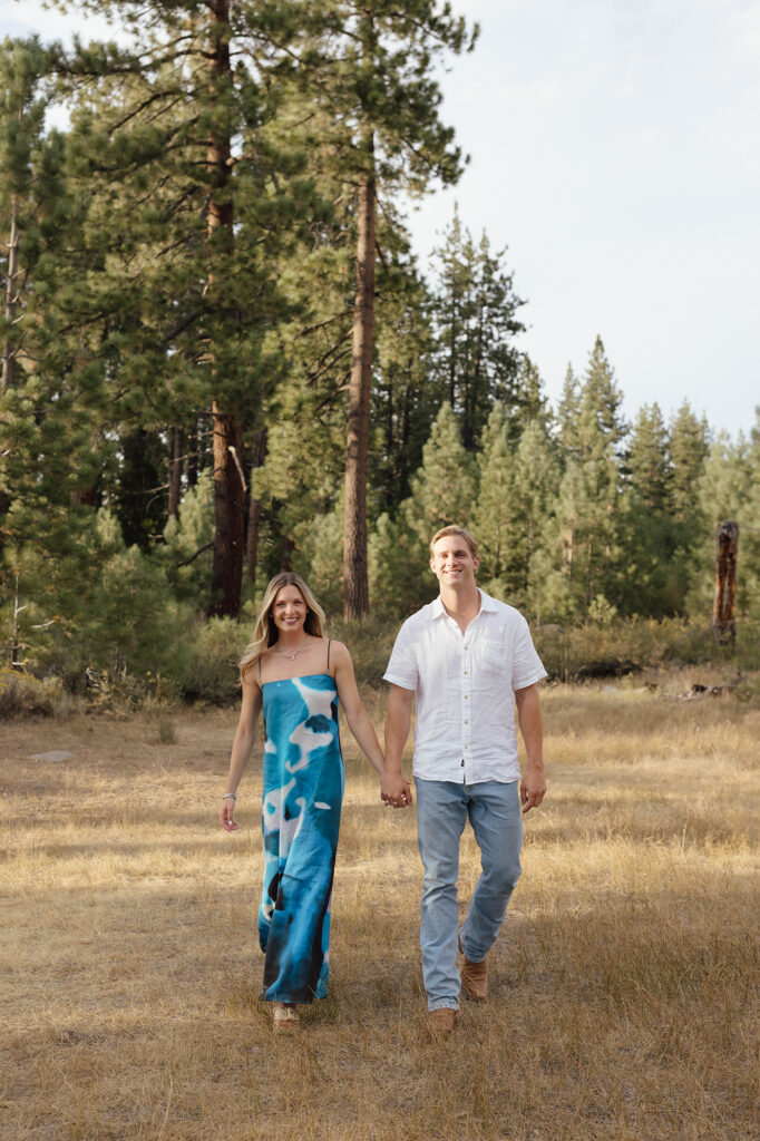 Newly engaged couple smiling and walking toward the camera through a grassy clearing surrounded by tall pines