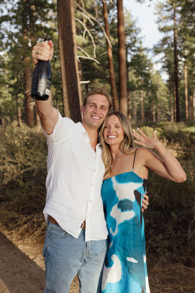 Smiling couple celebrating among pine trees, with the man raising a wine bottle and the woman showing her engagement ring